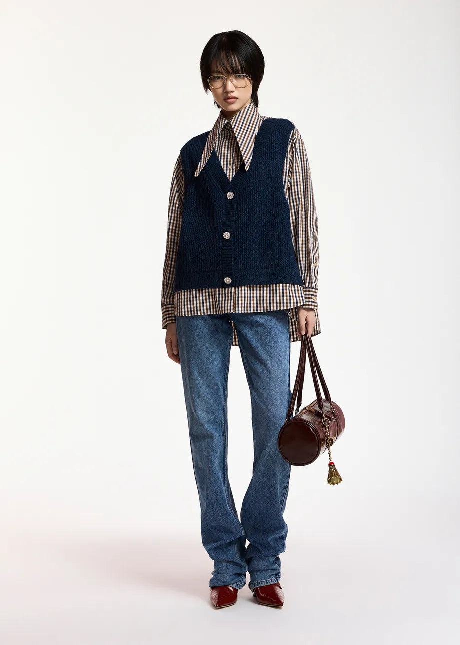A navy blue knit vest layered over a beige, brown, and white gingham shirt, featuring a pointed collar and three silver buttons on the front, set against a plain light gray background with a model wearing blue jeans and holding a purse.