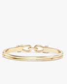 A gold bangle bracelet with a double-band design and a central horsebit clasp, photographed on a clean, light off-white background.