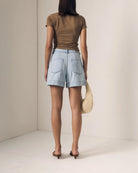 Person wearing a brown top and light blue shorts holding a woven bag against a plain background