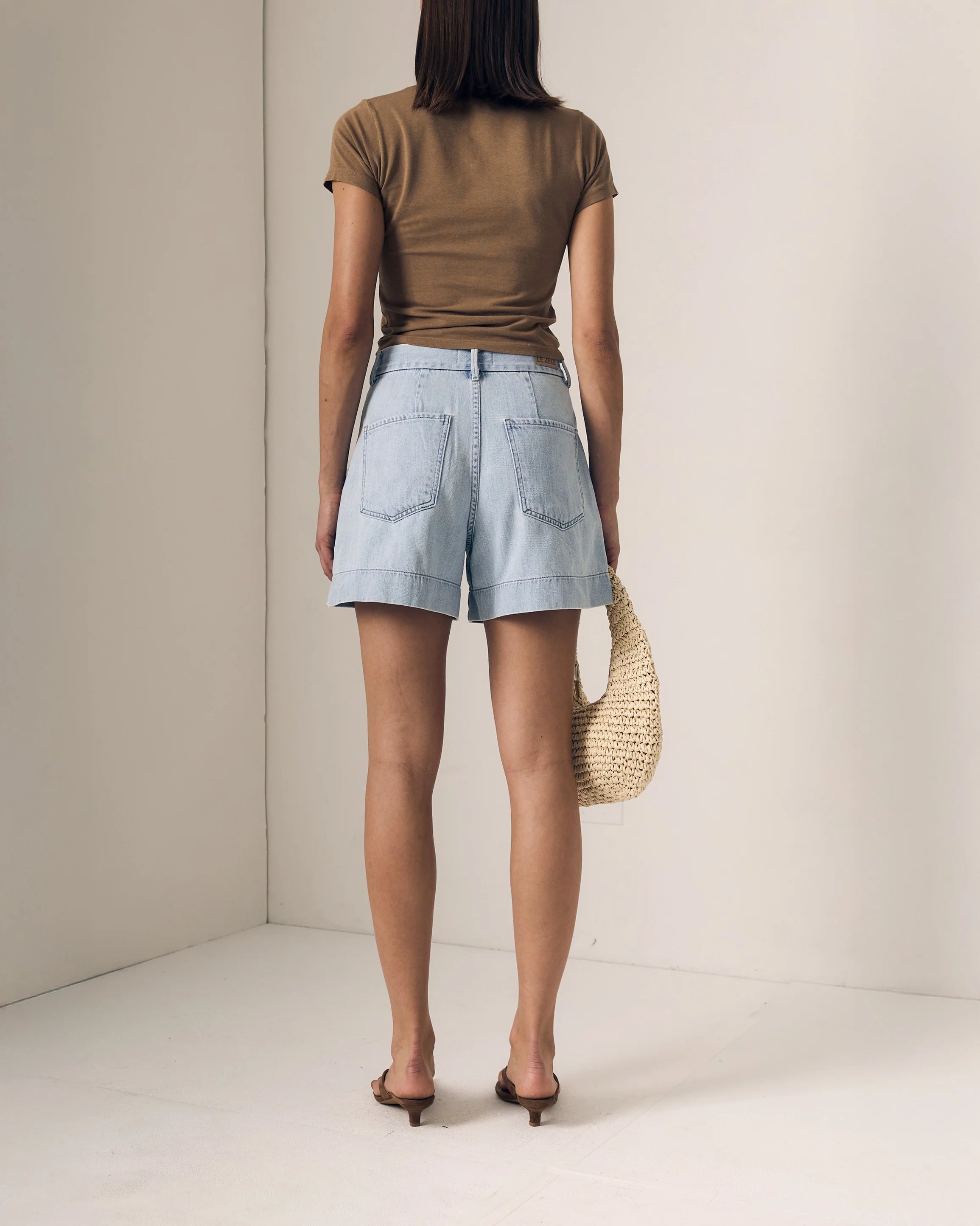 Person wearing a brown top and light blue shorts holding a woven bag against a plain background