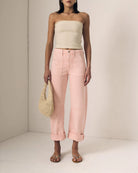 Woman wearing a beige strapless top and pink pants, holding a straw bag against a neutral background