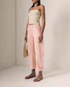 Woman wearing a beige strapless top and pink pants, holding a straw bag on a plain background