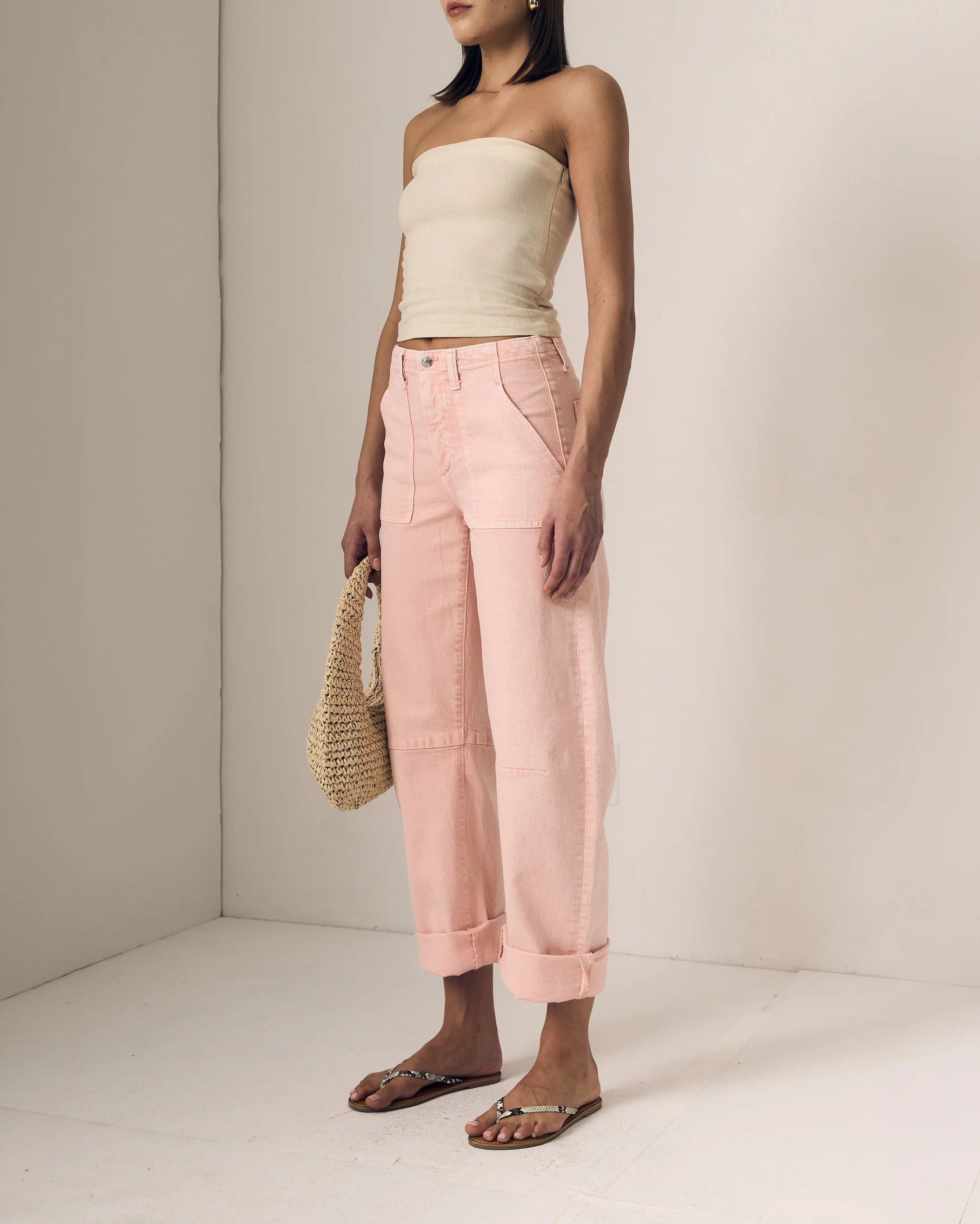 Woman wearing a beige strapless top and pink pants, holding a straw bag on a plain background