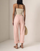 Woman wearing a strapless top and pink pants holding a straw bag against a neutral background