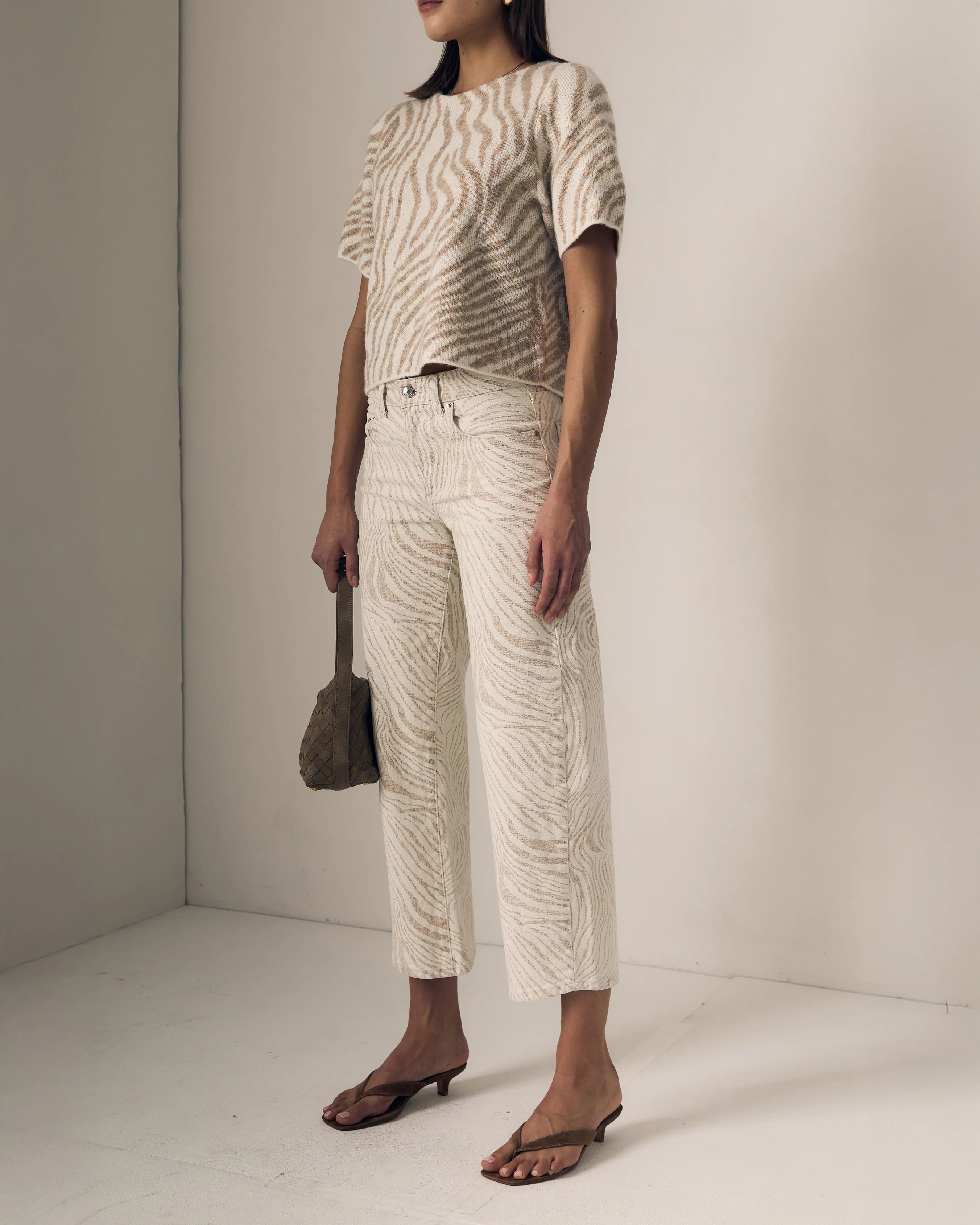 Person wearing a patterned top and white pants, holding a brown bag against a plain background