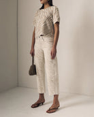Person wearing a patterned top and white pants, holding a brown bag against a plain background