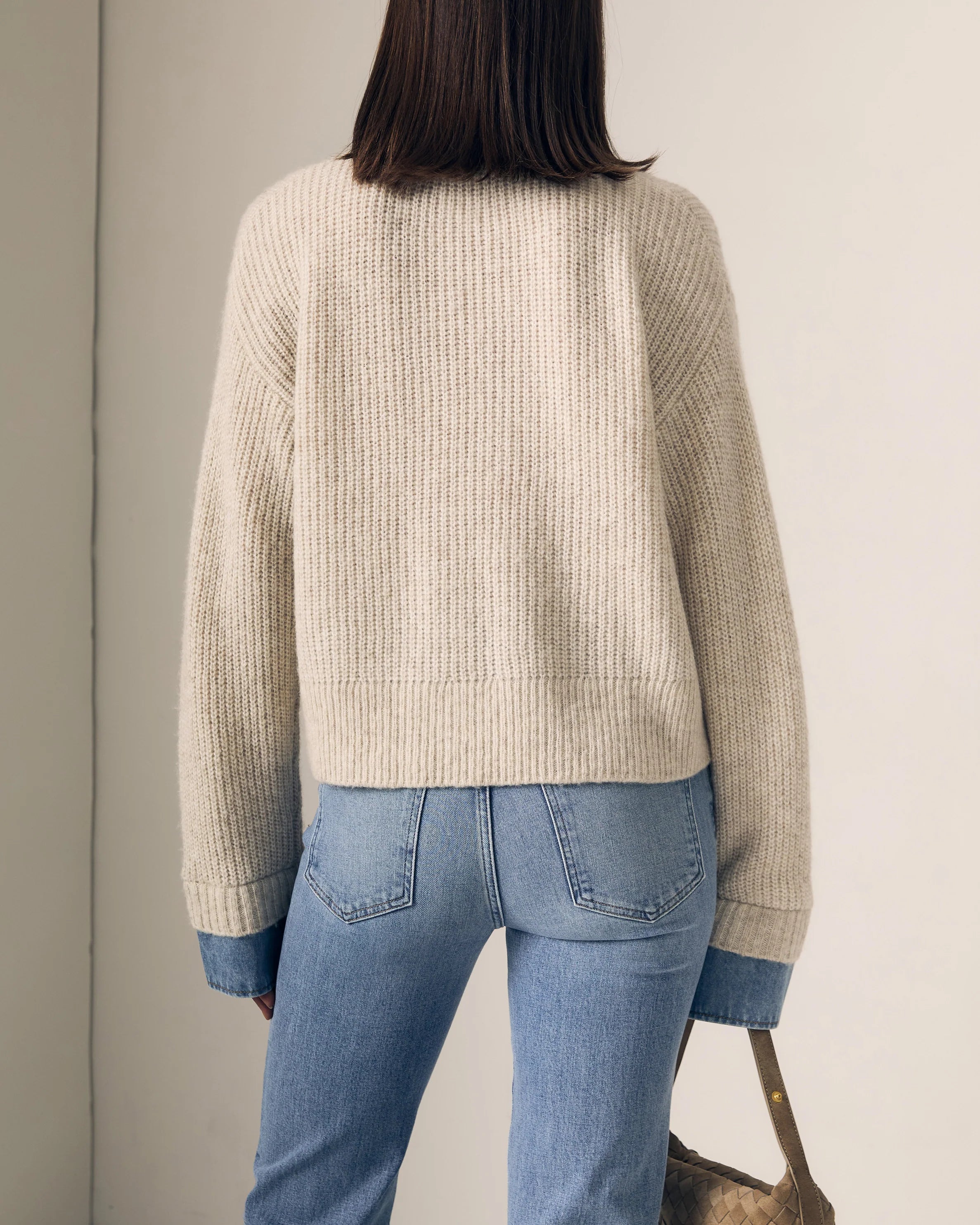 Person wearing a beige sweater and blue jeans against a neutral background
