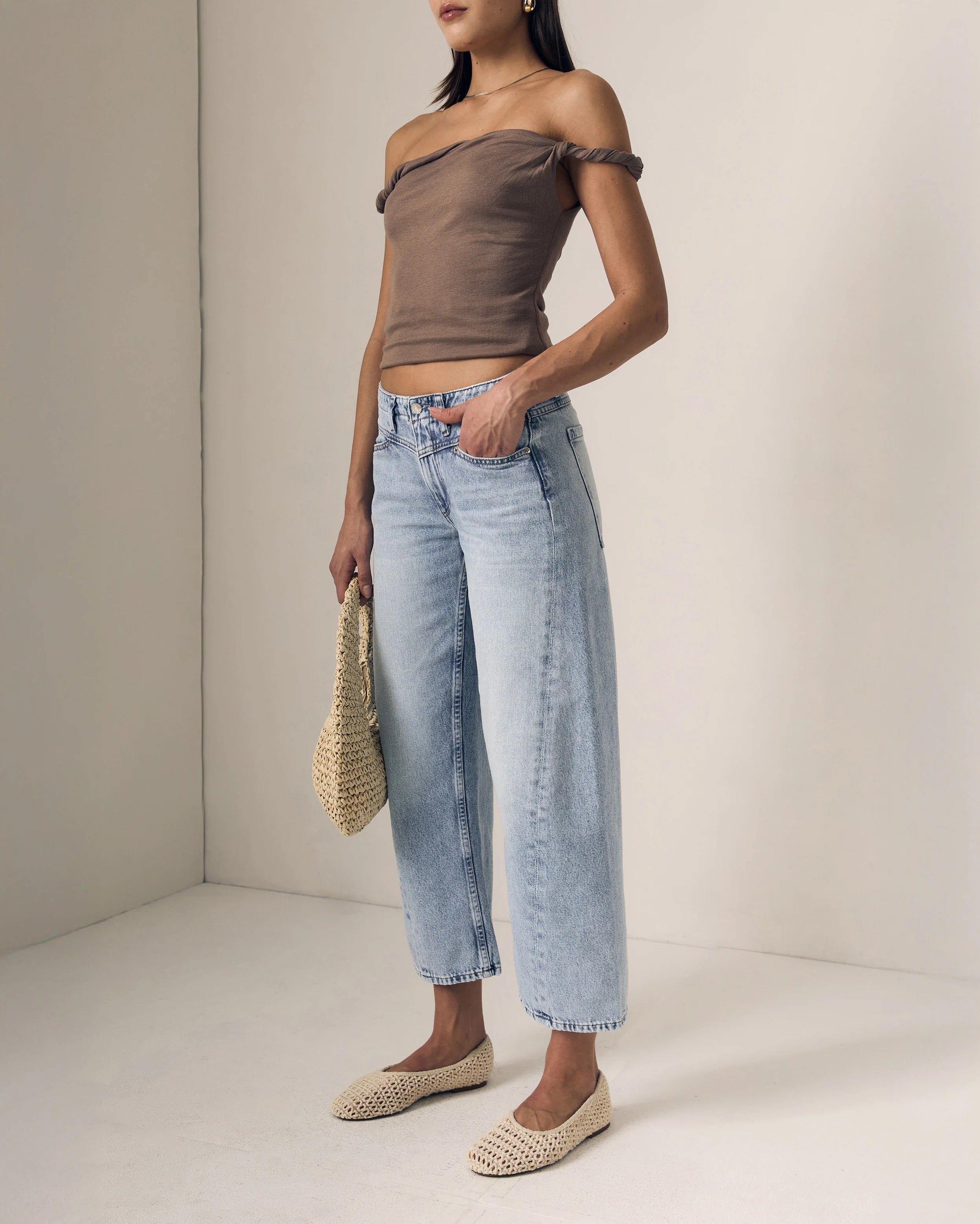 Woman wearing a brown off-shoulder top and light blue jeans, holding a straw bag on a plain background.