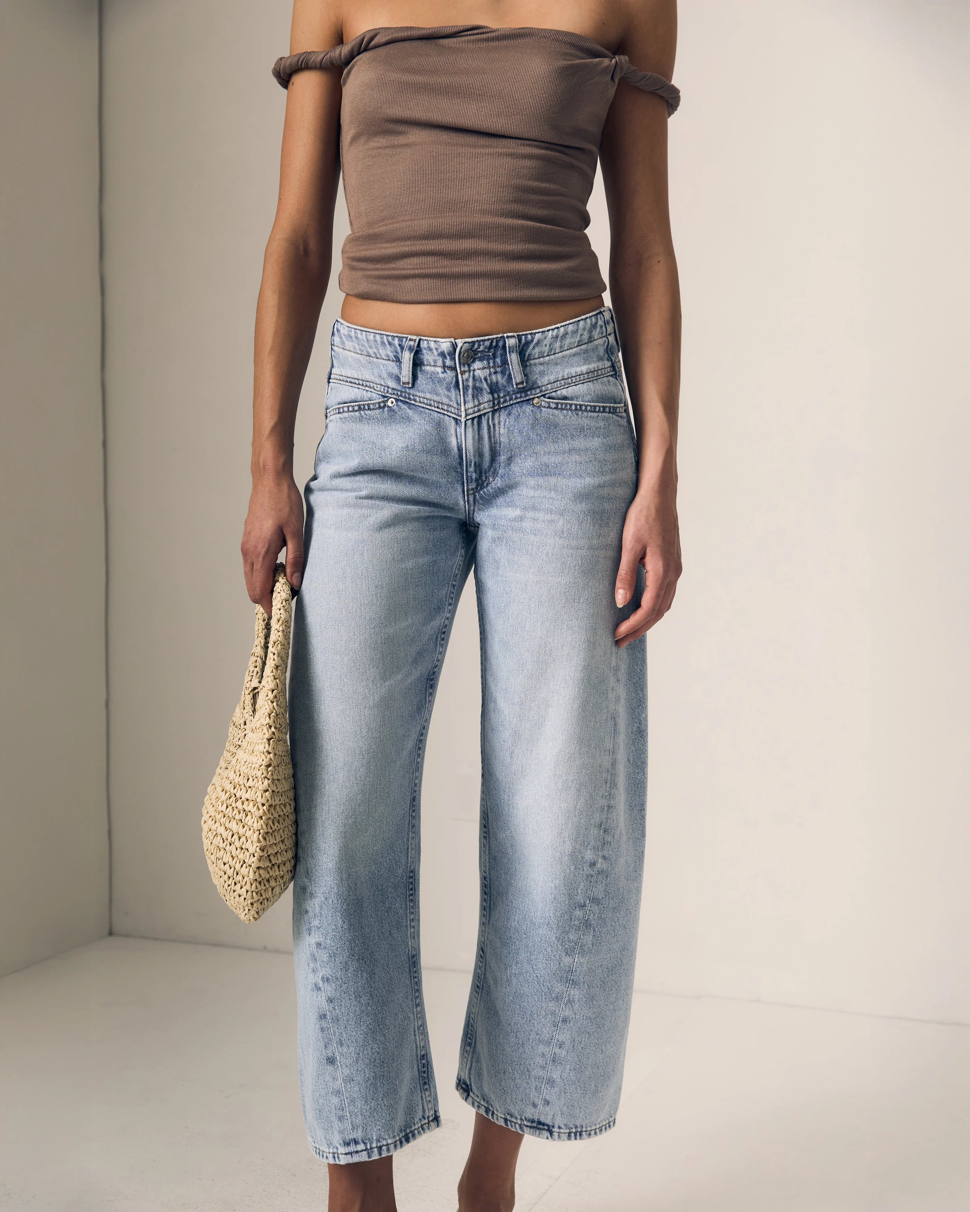 Person wearing a brown off-shoulder top and light blue jeans, holding a straw bag against a neutral background.