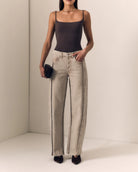 A model wearing a dark brown camisole top and light beige wide-leg jeans with black side stripes, holding a small black clutch against a minimalist beige background.