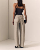 A woman shown from the back wearing light beige wide-leg jeans, a black spaghetti-strap camisole, and black heels in a minimalist studio setting.