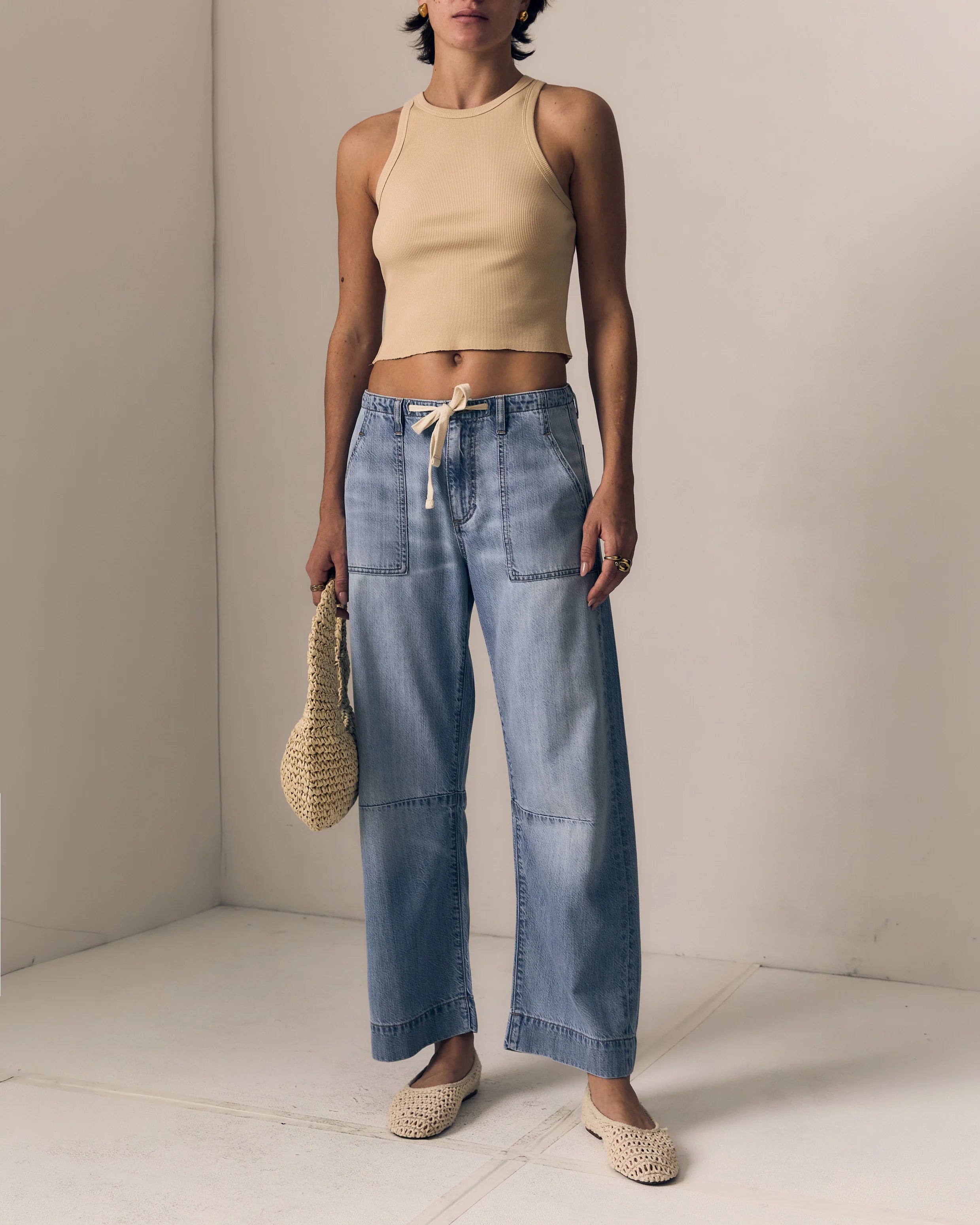 Woman wearing a beige top and blue jeans holding a woven bag against a plain background
