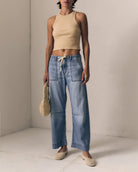 Woman wearing a beige top and blue jeans holding a woven bag against a plain background