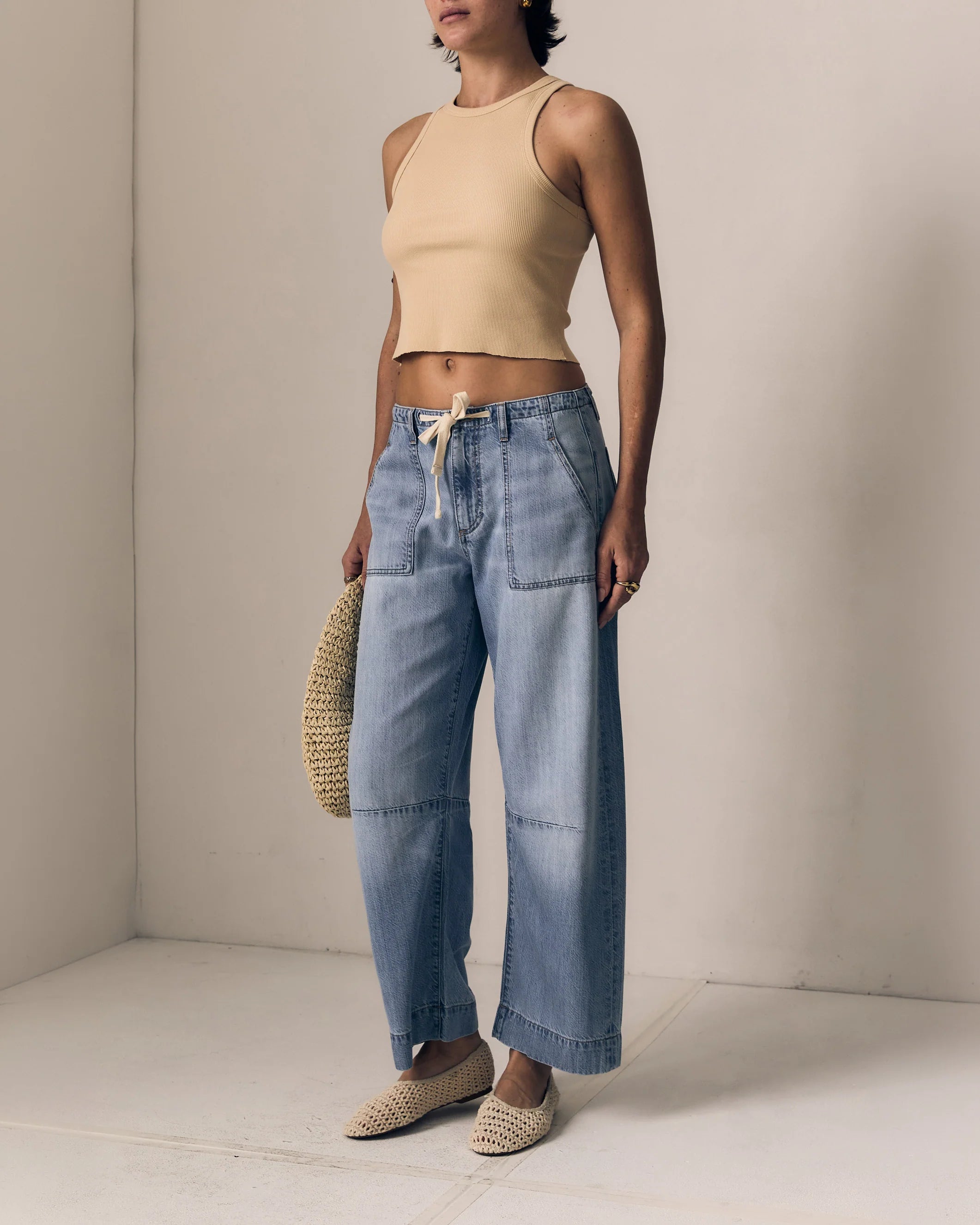 Woman wearing a beige top and blue jeans holding a straw hat against a neutral background