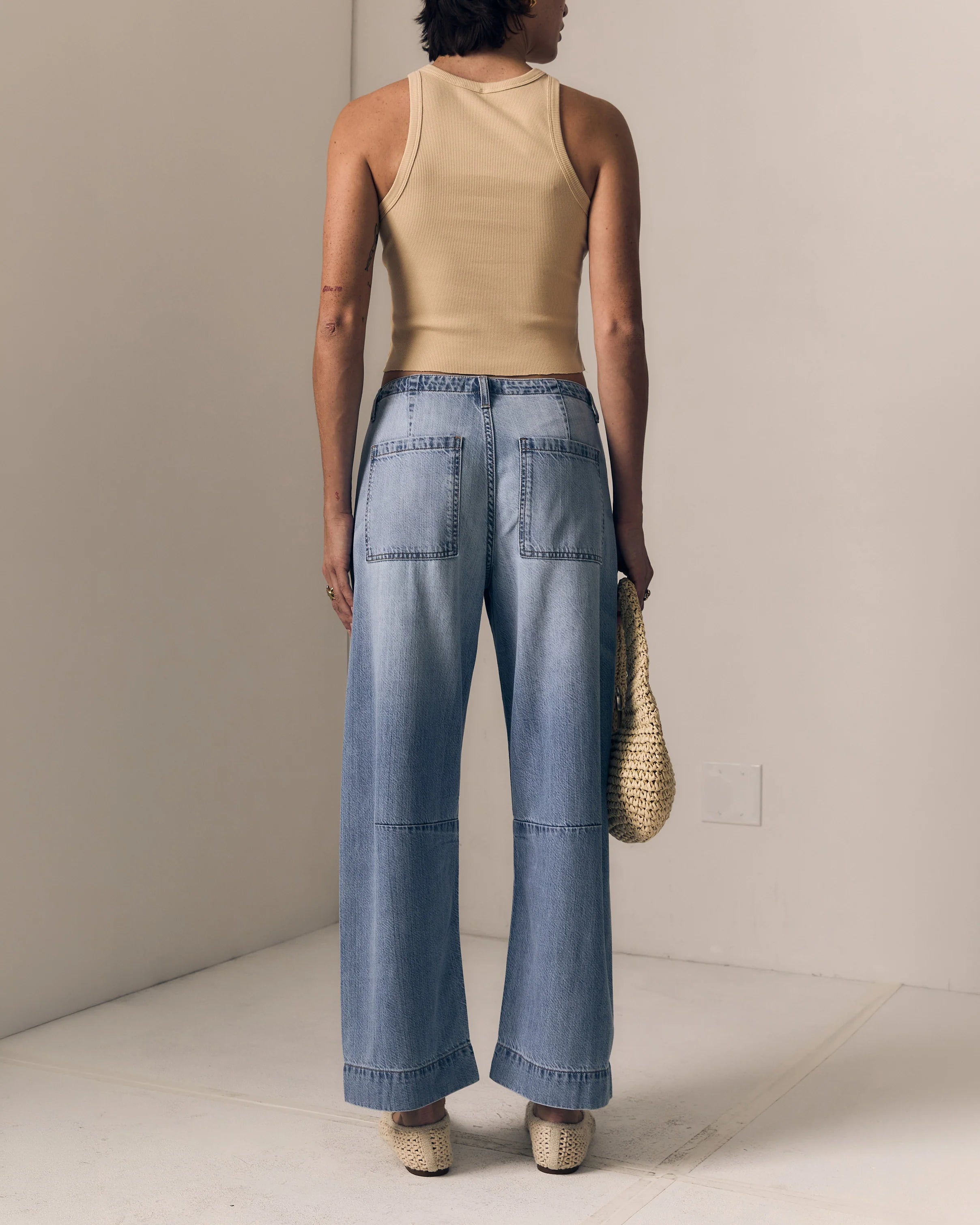 Person wearing a beige tank top and blue jeans, holding a straw bag, against a plain background.