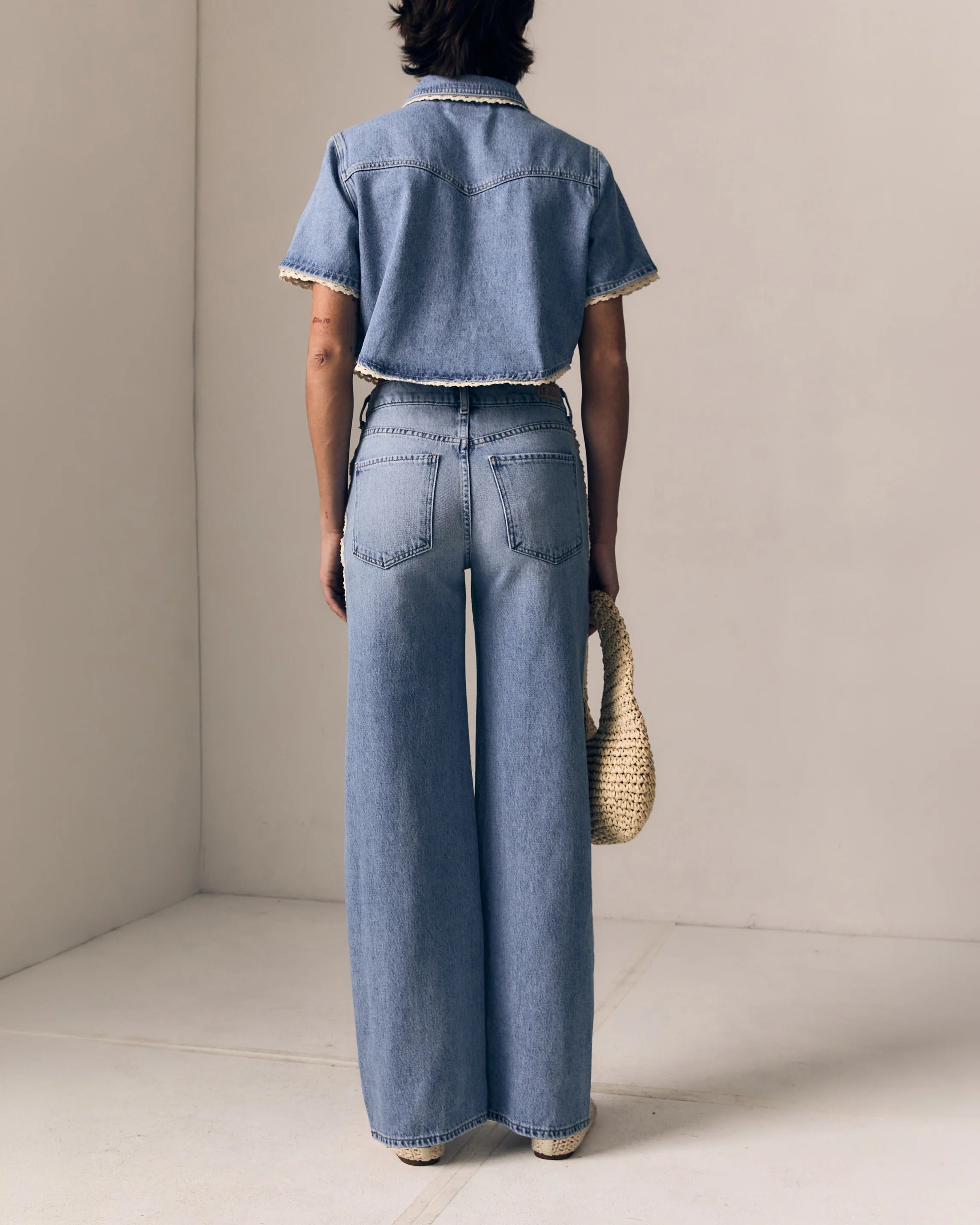 Person wearing a denim outfit with wide-leg jeans and a denim shirt, holding a woven bag.