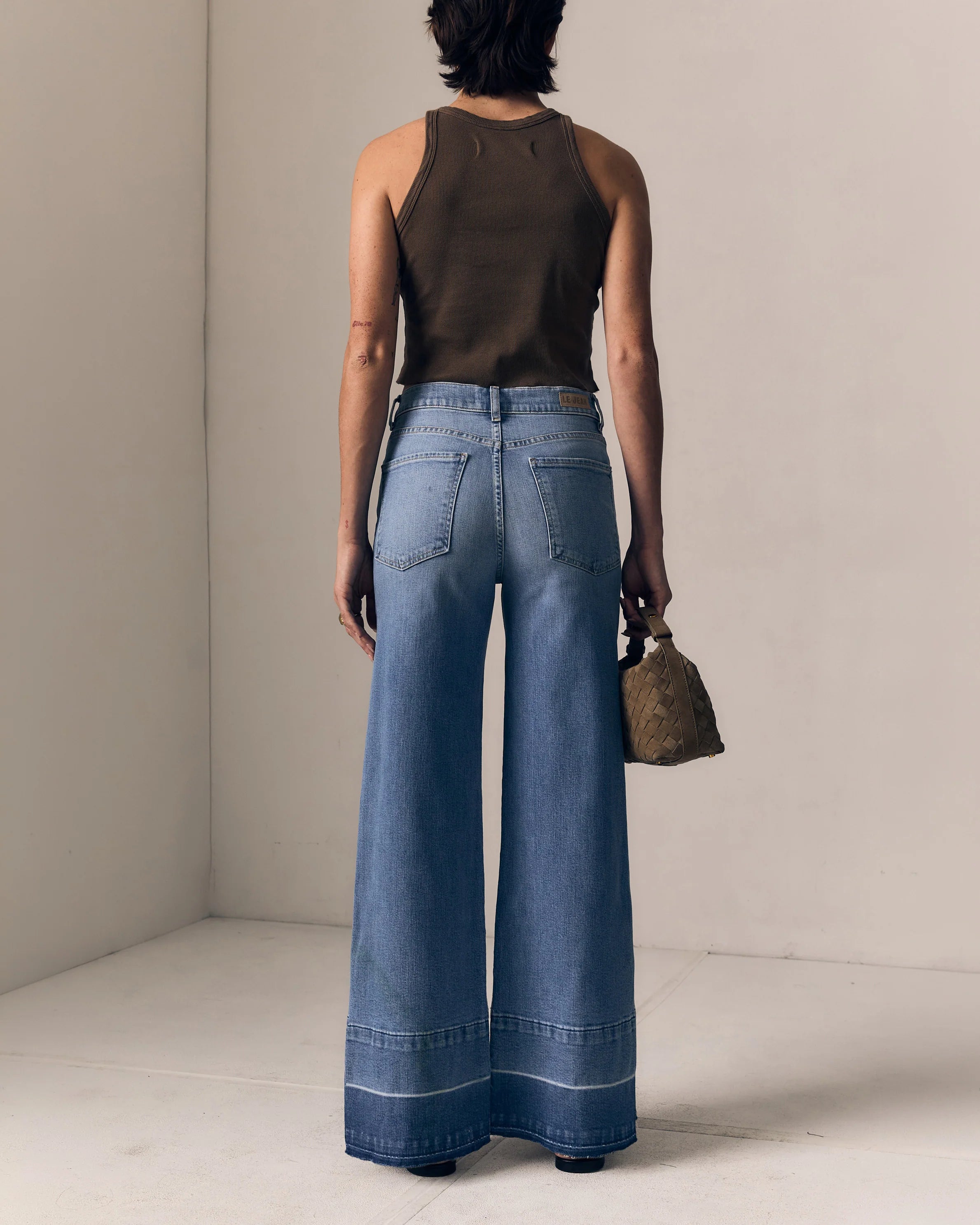Person wearing blue wide-leg jeans and a brown top, holding a small bag, against a neutral background.