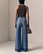 Person wearing blue wide-leg jeans and a brown top, holding a small bag, against a neutral background.