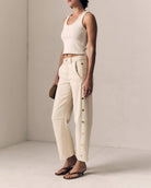 Woman wearing a white sleeveless top and beige pants against a plain background