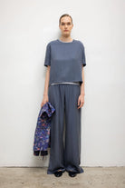 A model stands against a plain white wall wearing a loose short-sleeve blue-gray top and matching wide-leg pants, holding a patterned blue and purple jacket in one hand.