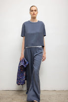 A model stands against a plain light gray wall wearing a loose short-sleeve blue-gray top and matching wide-leg pants, while holding a patterned blue jacket in one hand.