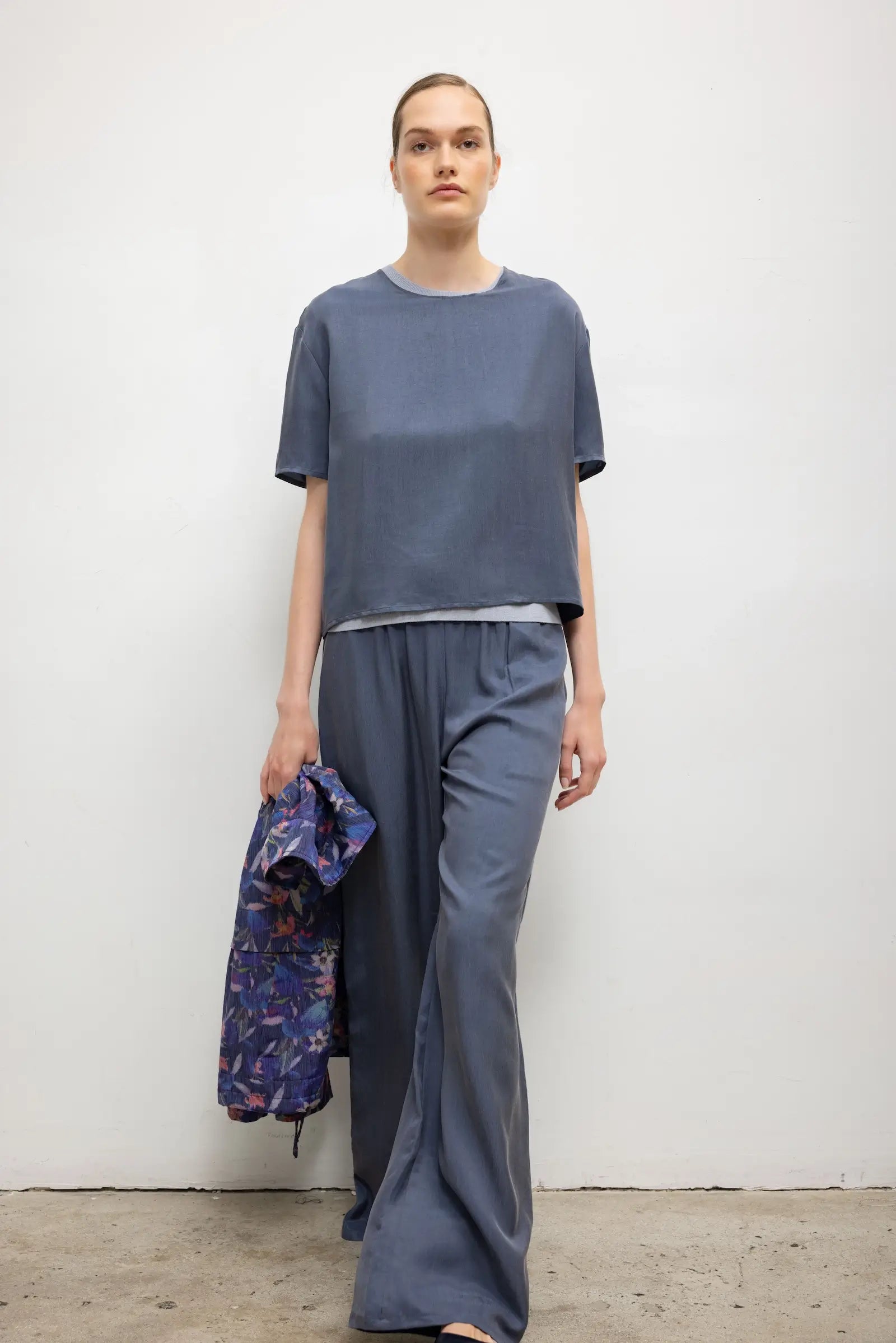 A model stands against a plain light gray wall wearing a loose short-sleeve blue-gray top and matching wide-leg pants, while holding a patterned blue jacket in one hand.