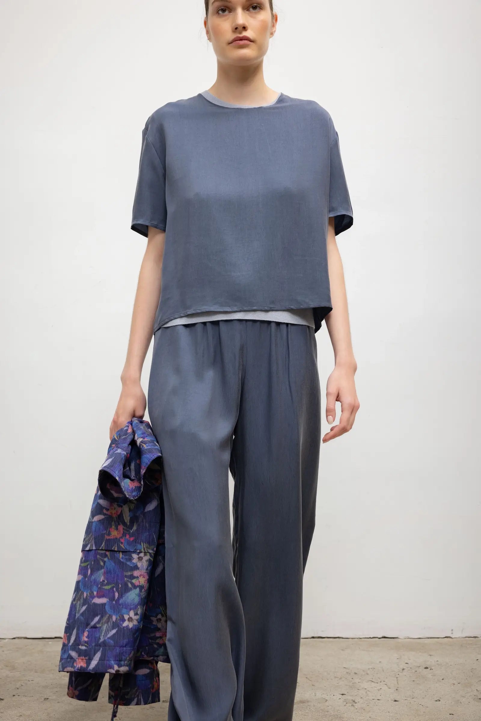 A model stands against a plain off-white wall wearing a loose short-sleeve blue-gray top and matching wide-leg pants, holding a folded floral-patterned garment in one hand.