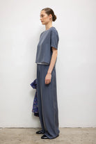 A woman stands in profile wearing a loose, short-sleeve gray-blue top and matching wide-leg pants, photographed against a plain off-white wall with a concrete floor.