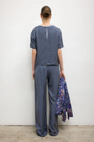 A model stands with their back to the camera wearing a slate blue short-sleeve top and matching wide-leg trousers, holding a purple floral patterned jacket in one hand against a plain white wall.