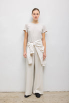 A model wearing a minimalist cream-colored outfit consisting of a short-sleeve crewneck t-shirt and matching wide-leg trousers, styled with a matching cream shirt tied around the waist.