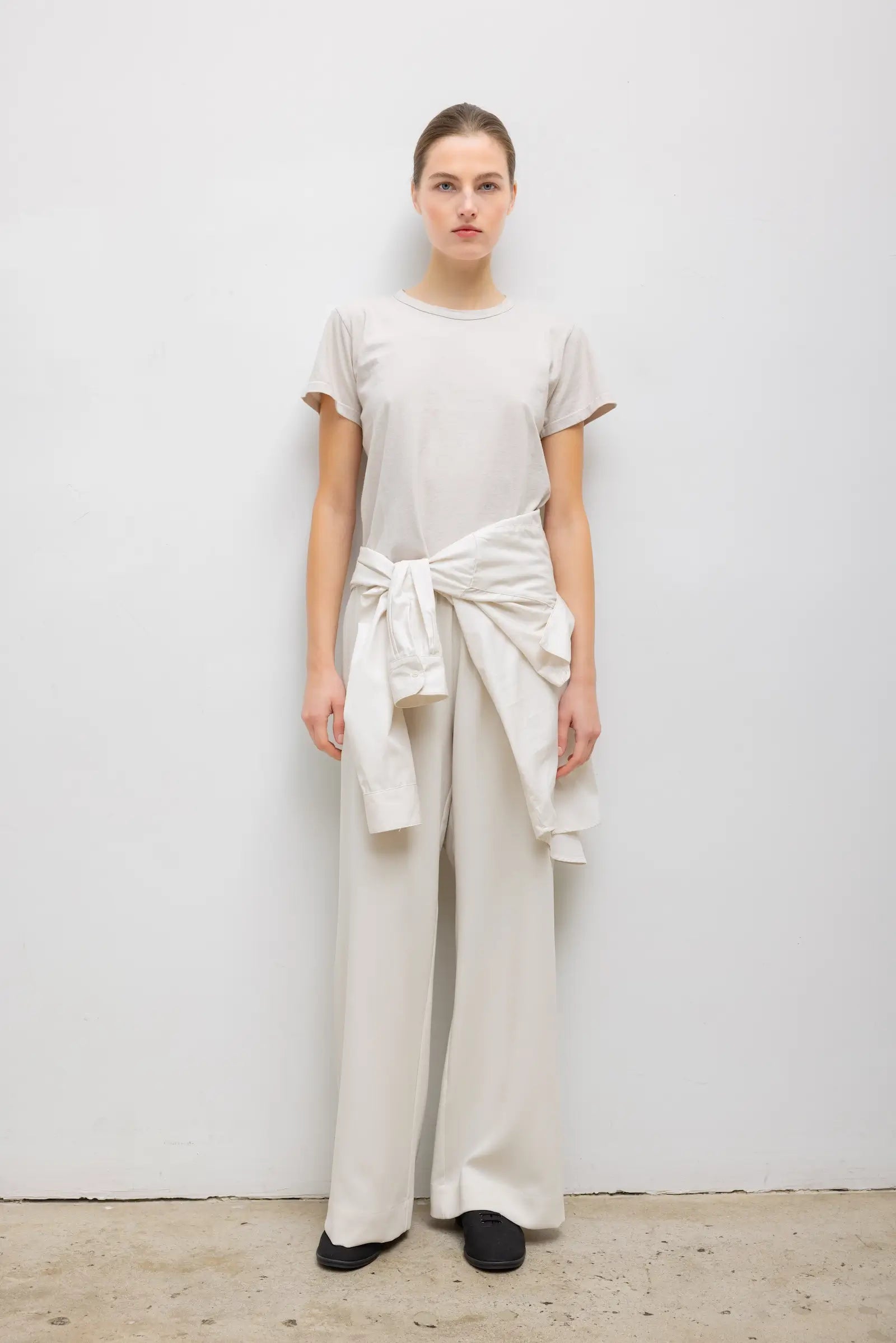 A model wearing a minimalist cream-colored outfit consisting of a short-sleeve crewneck t-shirt and matching wide-leg trousers, styled with a matching cream shirt tied around the waist.