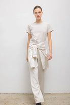 A model stands against a plain white wall wearing a light beige short-sleeve t-shirt, matching wide-leg trousers, and a light beige shirt tied around the waist.