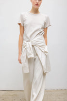 A model wearing a minimalist cream-colored outfit consisting of a short-sleeve crewneck t-shirt and matching wide-leg trousers, styled with a matching cream shirt tied around the waist.