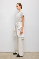 A model wearing a minimalist cream-colored outfit consisting of a short-sleeve crewneck t-shirt and matching wide-leg trousers, styled with a matching cream shirt tied around the waist.