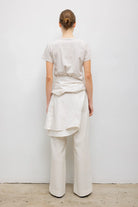A model wearing a minimalist cream-colored outfit consisting of a short-sleeve crewneck t-shirt and matching wide-leg trousers, styled with a matching cream shirt tied around the waist from a back side view.