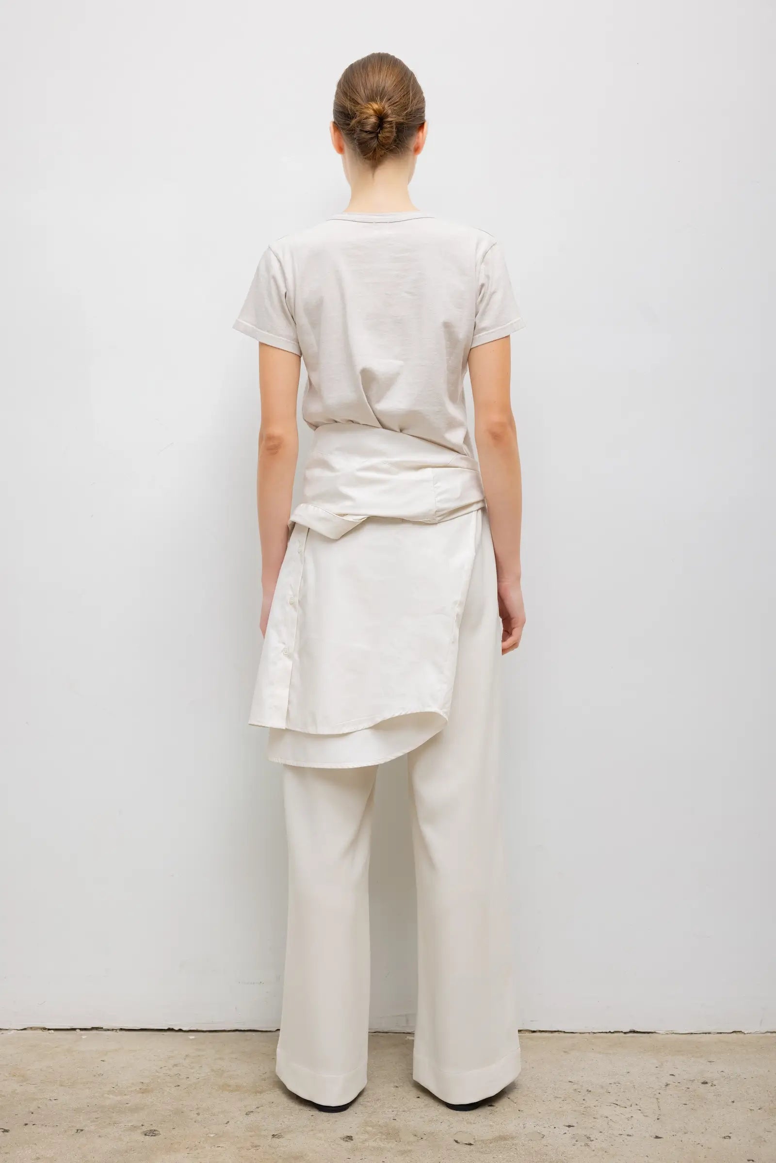 A model wearing a minimalist cream-colored outfit consisting of a short-sleeve crewneck t-shirt and matching wide-leg trousers, styled with a matching cream shirt tied around the waist from a back side view.