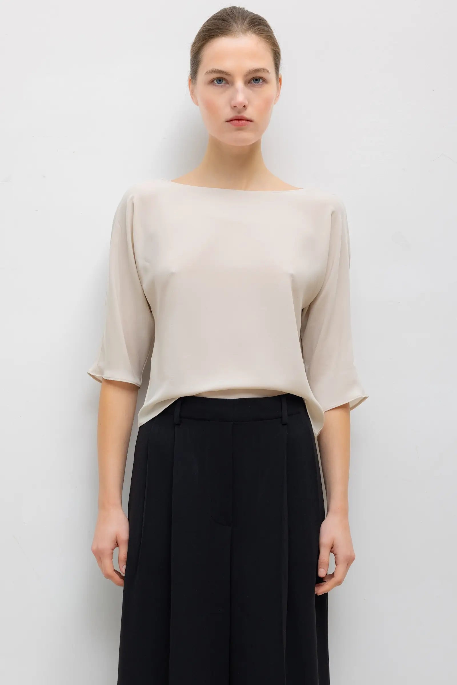 A woman modeling a light beige boat-neck top with three-quarter sleeves and black wide-leg trousers, standing against a plain light gray wall.