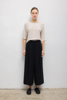 A model stands against a plain light gray wall wearing a beige short-sleeve top and black wide-leg cropped pants with black slip-on shoes.