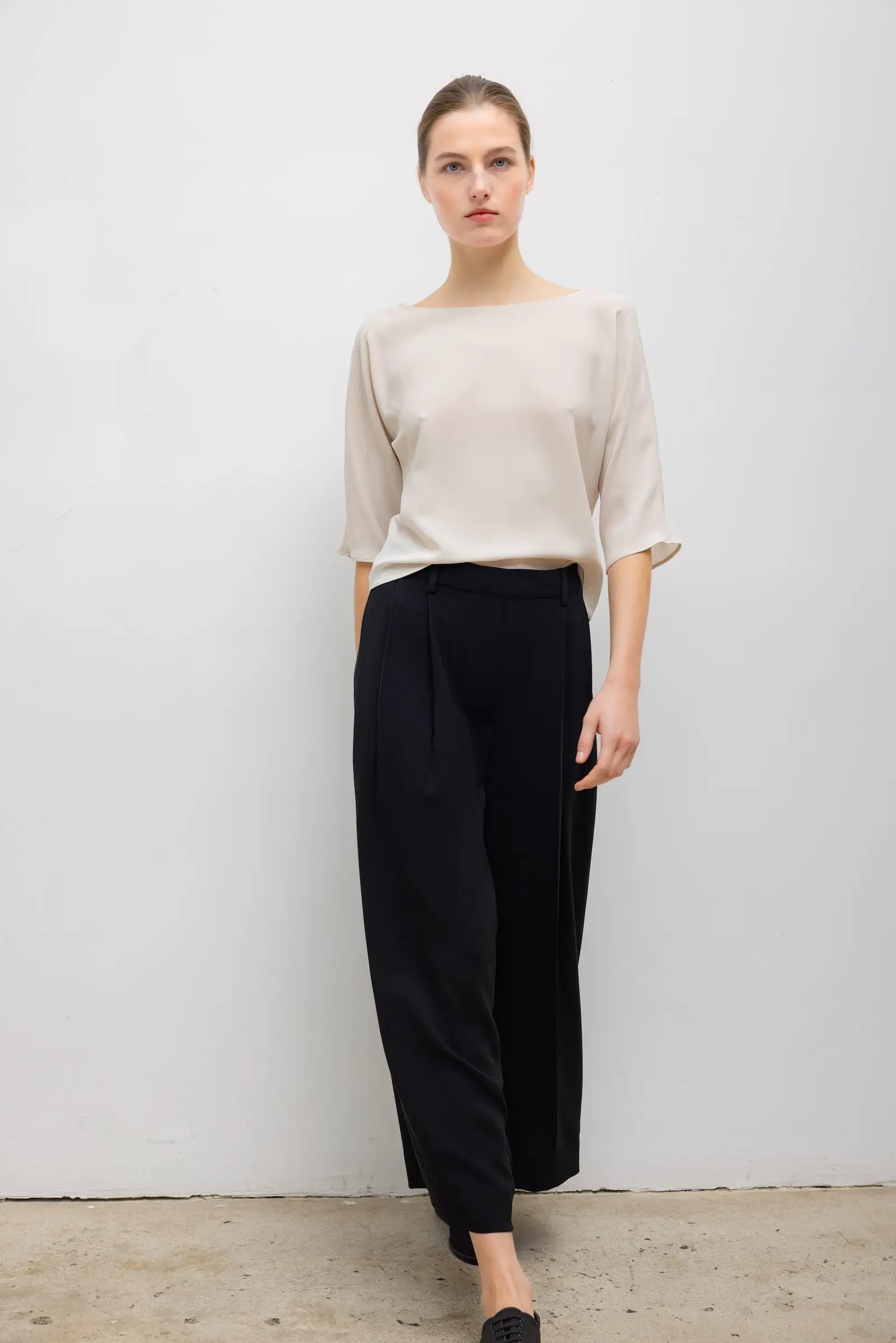A woman modeling a light beige short-sleeve top and black wide-leg cropped trousers against a plain light gray wall.
