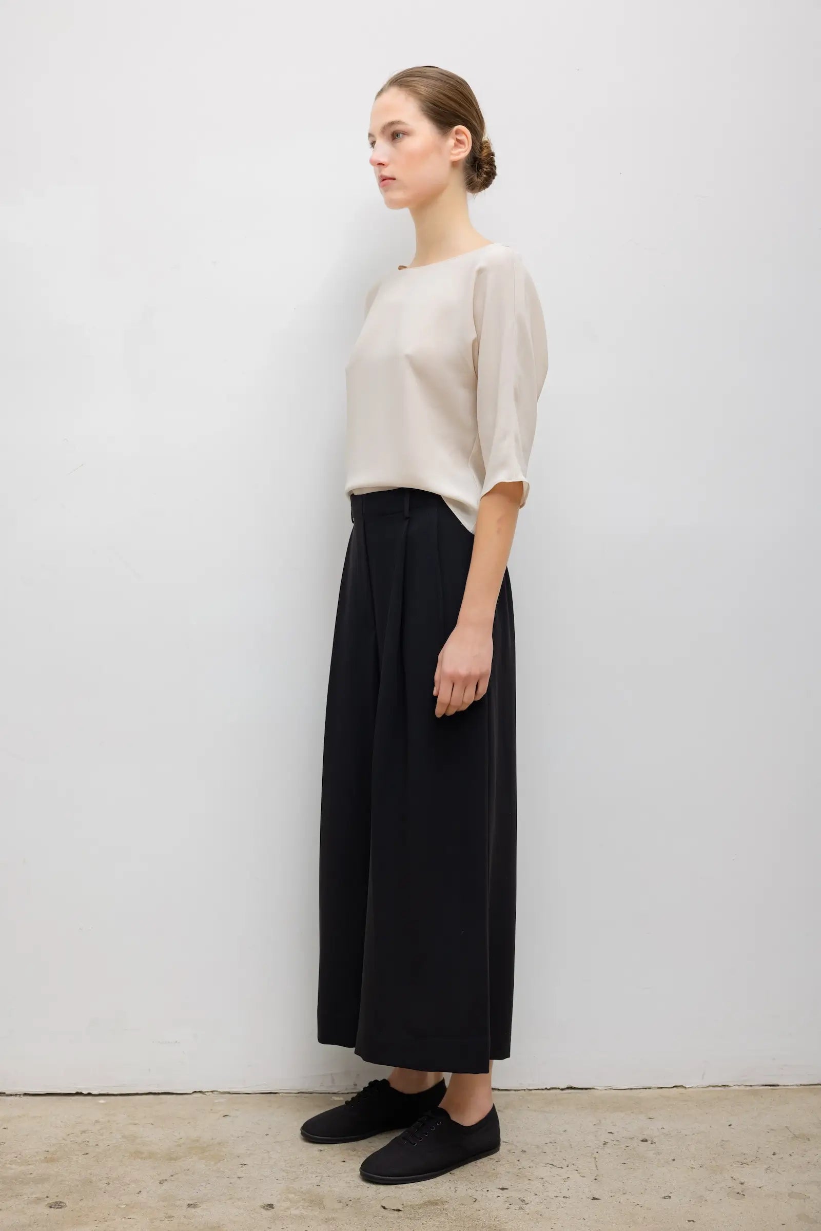 A woman stands in profile wearing a light beige short-sleeve top and black wide-leg cropped pants with black slip-on shoes, photographed against a plain light gray wall.