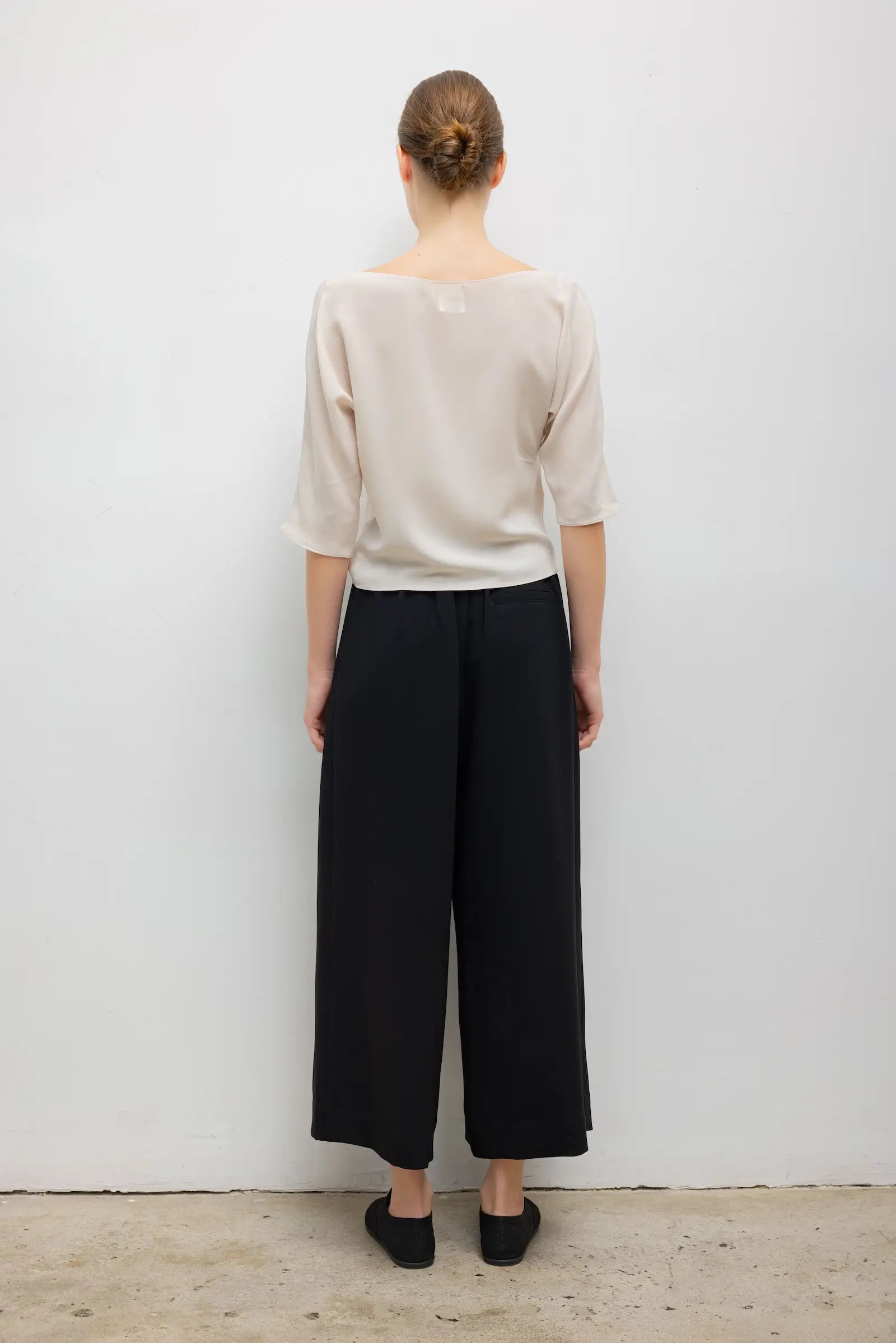 A woman shown from the back wearing a light beige short-sleeve top and black wide-leg cropped pants, standing against a plain white wall.