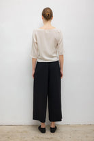 A woman shown from the back wearing a light beige short-sleeve top and black wide-leg cropped pants, standing against a plain white wall.