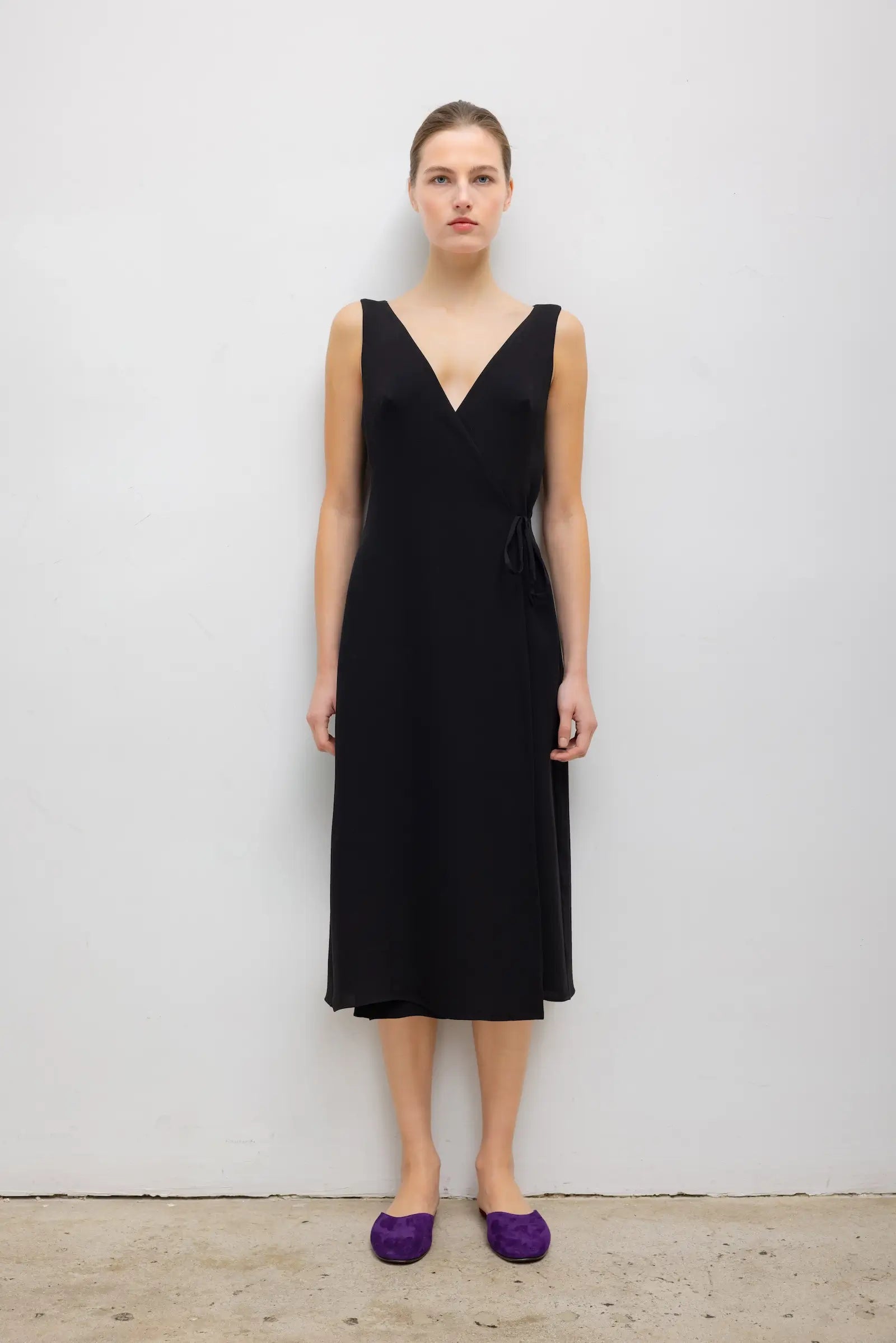 A woman stands against a plain light gray wall wearing a sleeveless black midi dress with a deep V-neck and purple flats.