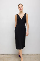 A woman modeling a sleeveless black midi dress with a wrap-style V-neck bodice and a side tie detail, standing against a plain light gray wall.