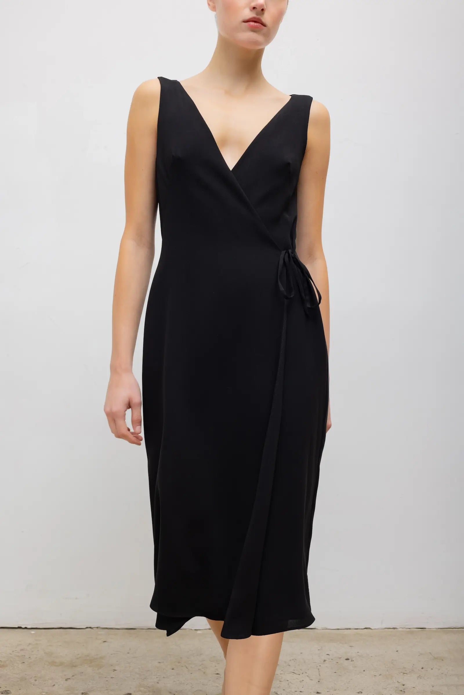 A woman wearing a sleeveless black wrap-style midi dress with a V-neckline and side tie detail, standing against a plain light gray wall.