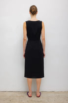 A woman viewed from the back wearing a sleeveless black knee-length dress, standing against a plain light gray wall on a concrete floor.