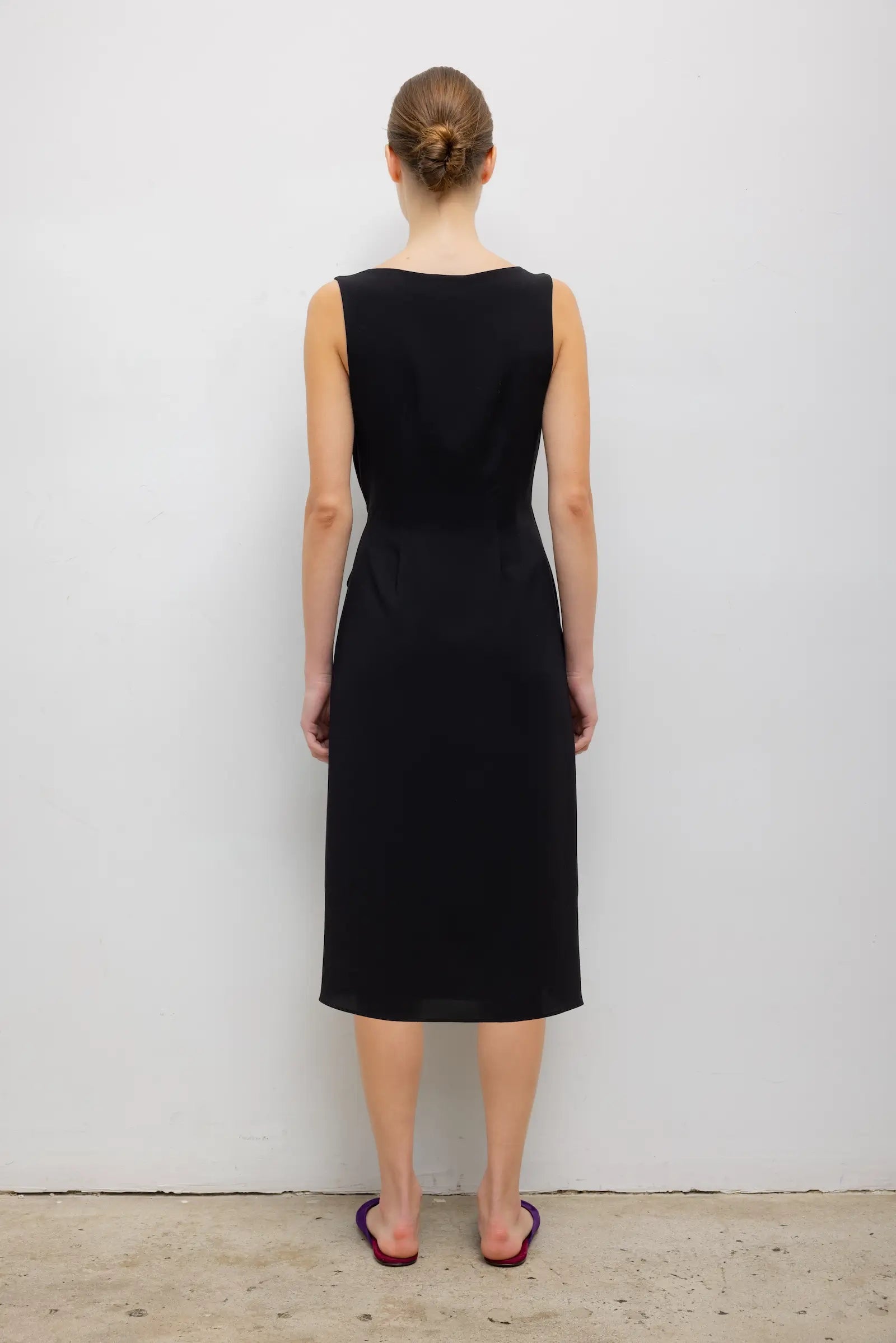 A woman viewed from the back wearing a sleeveless black knee-length dress, standing against a plain light gray wall on a concrete floor.