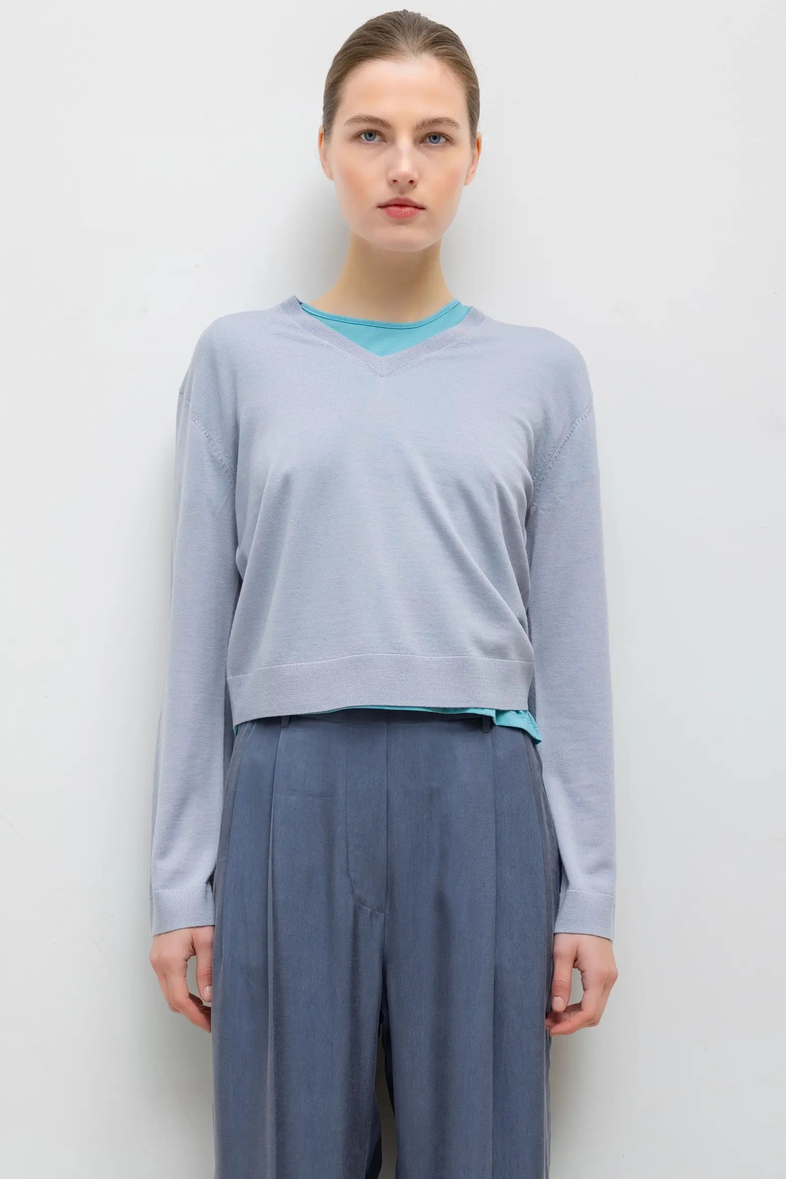 A woman modeling a light blue-gray long-sleeve cropped sweater with a bright turquoise inner collar and hem detail, paired with slate blue wide-leg trousers, against a plain light gray background.