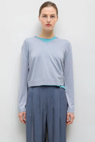 A woman modeling a light blue-gray long-sleeve cropped sweater with a bright turquoise inner collar and hem detail, paired with slate blue wide-leg trousers, against a plain light gray background.