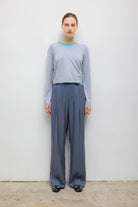 A model stands against a plain white wall wearing a light blue-gray long-sleeve crop top with a turquoise collar and matching wide-leg trousers in a muted blue-gray color.